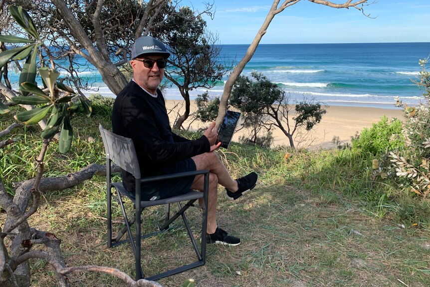 Peter Silburn sitting on a chair at from Yaroomba beach with a laptop.