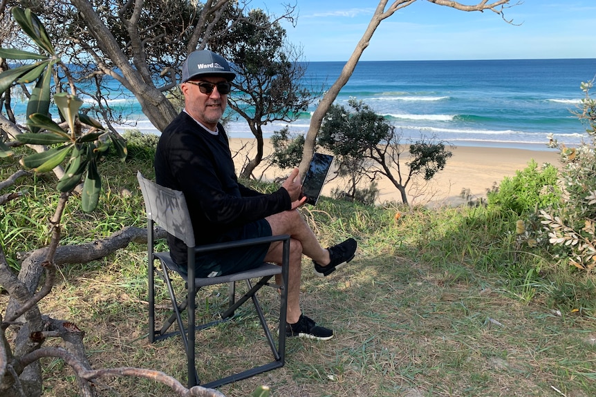 Peter Silburn sitting on a chair at from Yaroomba beach with a laptop.