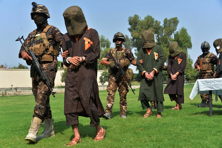Armed soldiers lead men who have bags over their heads across a lawn.