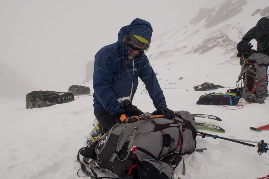 Huw Kingston packs a bag surrounded by snow with his skis on the ground next to him. He's wearing a blue jacket and beanie.