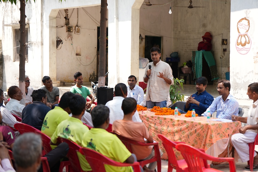 Ramveer Tanwar speaks to a group of people sitting on red plastic chairs.
