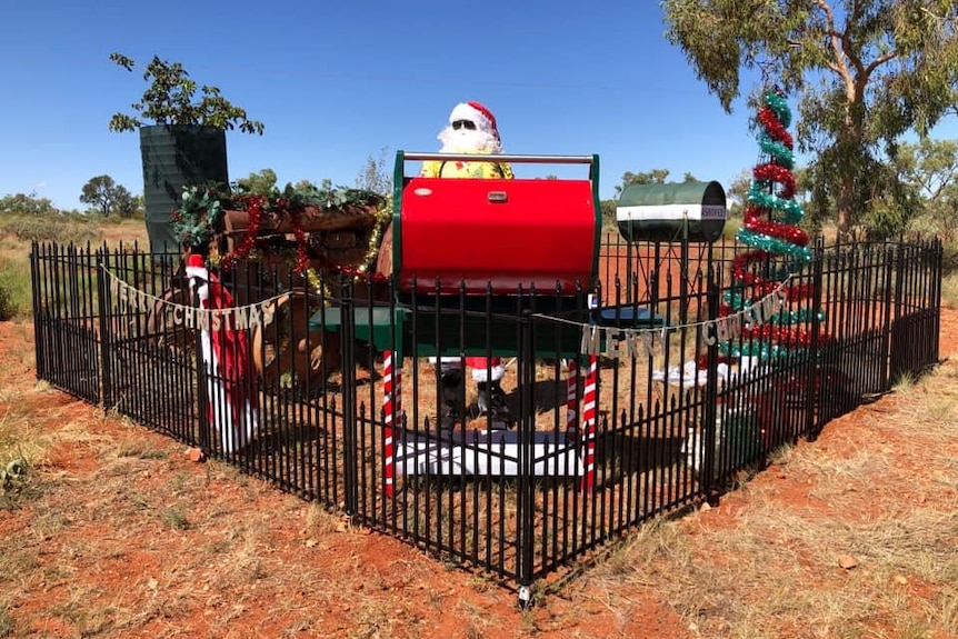A Santa standing behind a black fence with a red BBQ and Christmas tree.