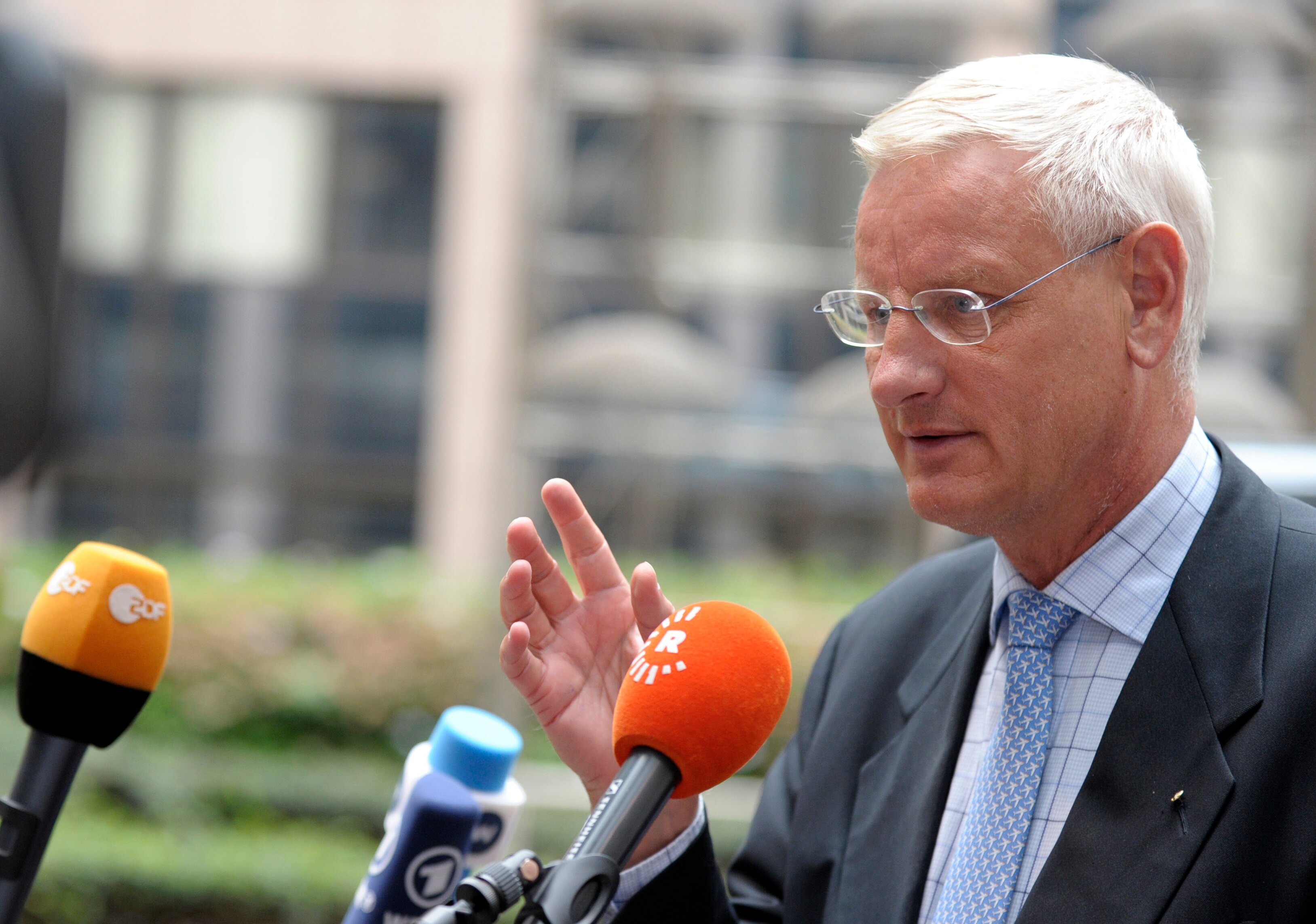 A man wearing a suit and tie speaks into microphones.