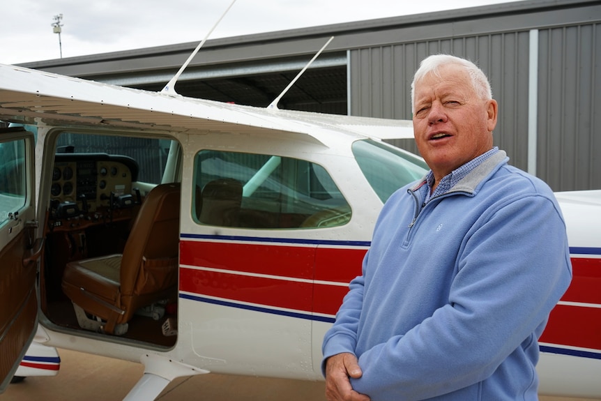 A man with white hair stands next to a red and white single-engine plane.