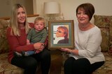 Three generations- grandmother, mother and baby son hold a framed photograph of a man.