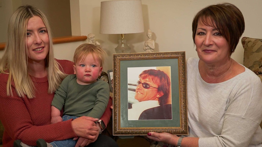 Three generations- grandmother, mother and baby son hold a framed photograph of a man.