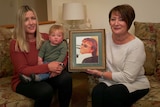 Three generations- grandmother, mother and baby son hold a framed photograph of a man.