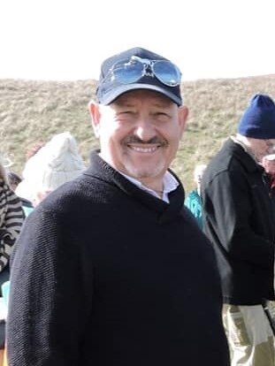 A smiling man in a baseball cap and jumper at an outdoor event.