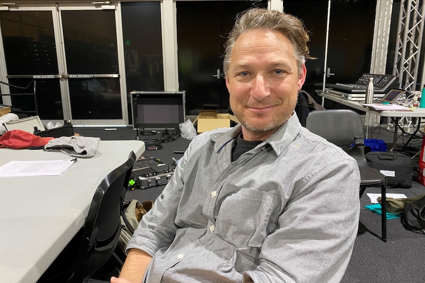 A man in a button-up grey shirt sits smiling at the camera.