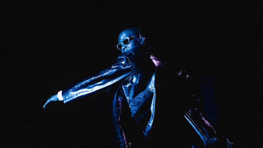 Nez stands in blue light wearing a black leather jacket and sunglasses. He's turned away from the camera. Black background.
