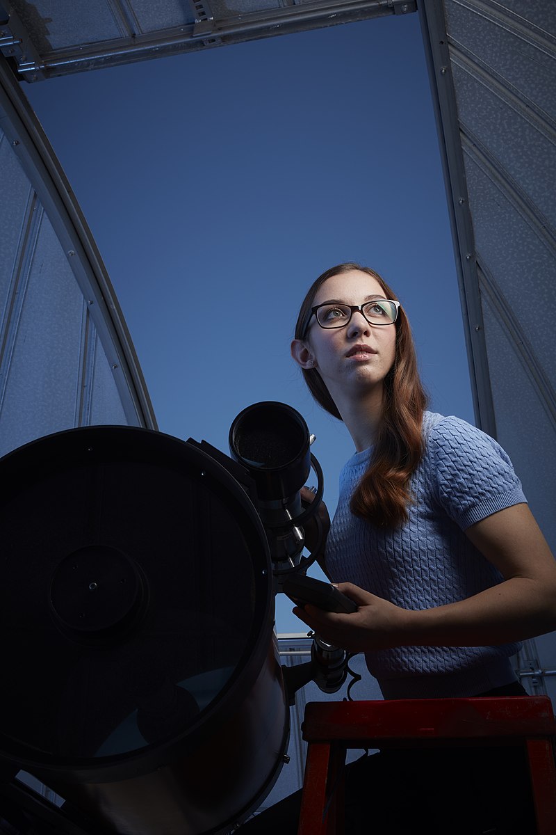 A young woman sits next to a telescope.