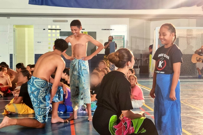 Two senior Samoan siva students mentor two younger students in a large school hall filled with kids. 