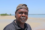 An middle-aged Indigenous man wearing a brown cap and a black shirt, standing on a beach.