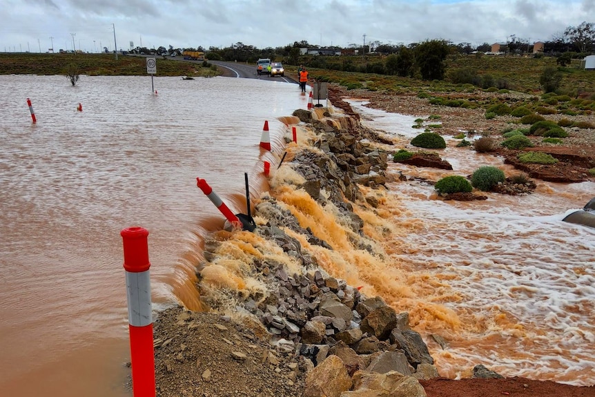 Flood water rushes over a road, with orange bollards on either side of the road marking the way