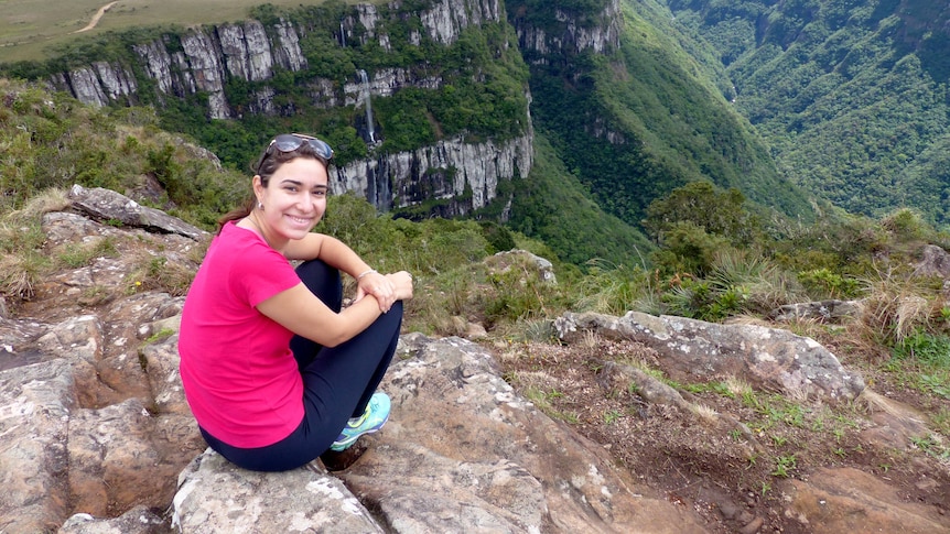 A young woman perches on a rock overlooking a valley.