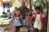 A man stands in a classroom surrounded by children and groceries.