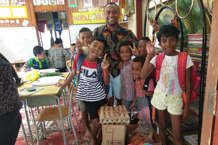 A man stands in a classroom surrounded by children and groceries.