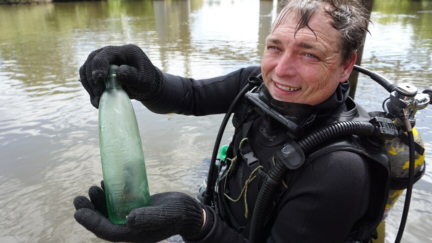 A diver stands in front of a river with a bottle he has found under the water