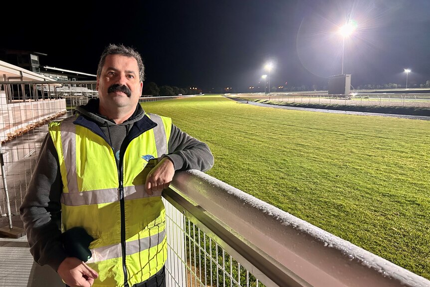A man in a high vis shirt stands next to a green grass racetrack under lights in the darkness