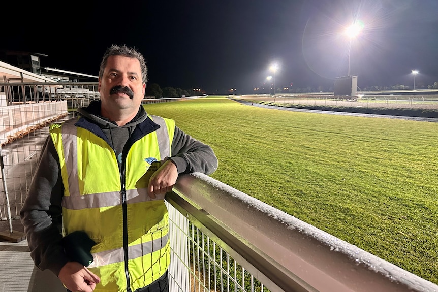 A man in a high vis shirt stands next to a green grass racetrack under lights in the darkness