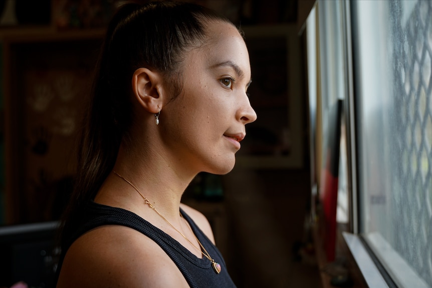 A slightly smiling young woman looking out a window, hair tied back, black tee-shirt.