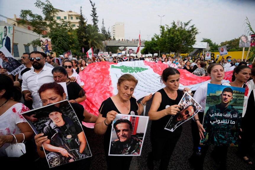 Crying women marching in front of a crowd holding a large Lebanese flag hold portraits of family members killed.