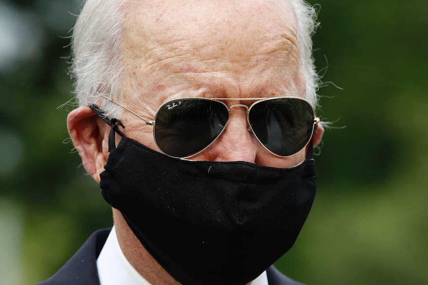 Grey-haired man with black sunglasses and facemask wearing a black suit and dark striped tie