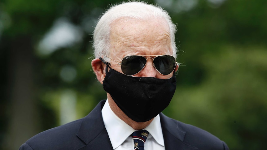 Grey-haired man with black sunglasses and facemask wearing a black suit and dark striped tie