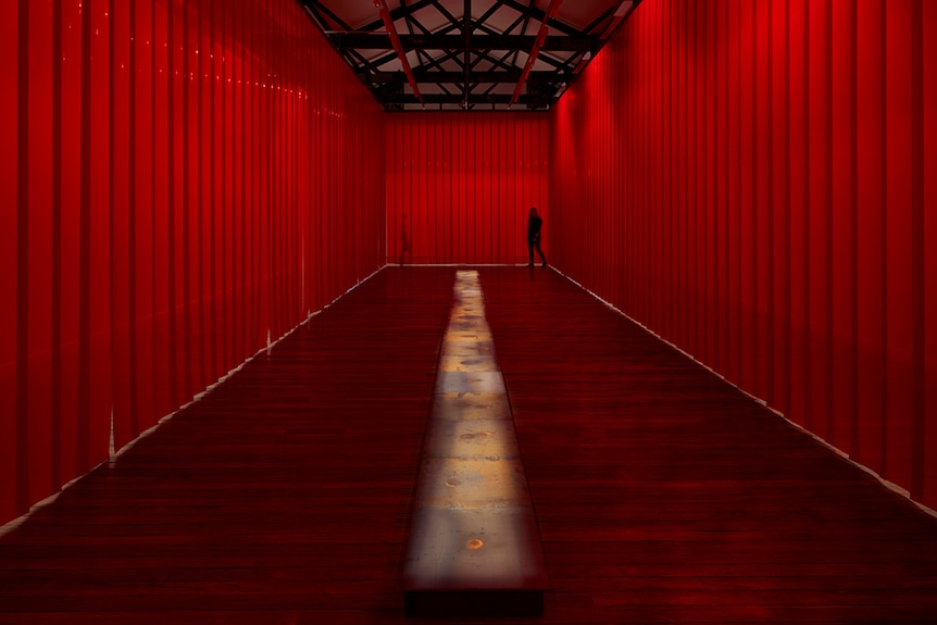 A figure stands in long narrow room lined from ceiling to floor with red curtains with long and low artwork at room's centre.