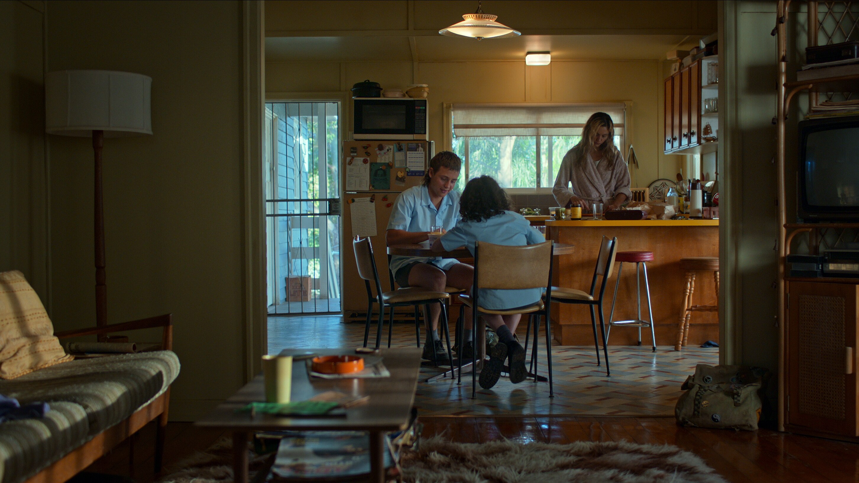 Two teenage boys sit at a retro kitchen table, with their mum behind them preparing food at the counter, a big window behind her