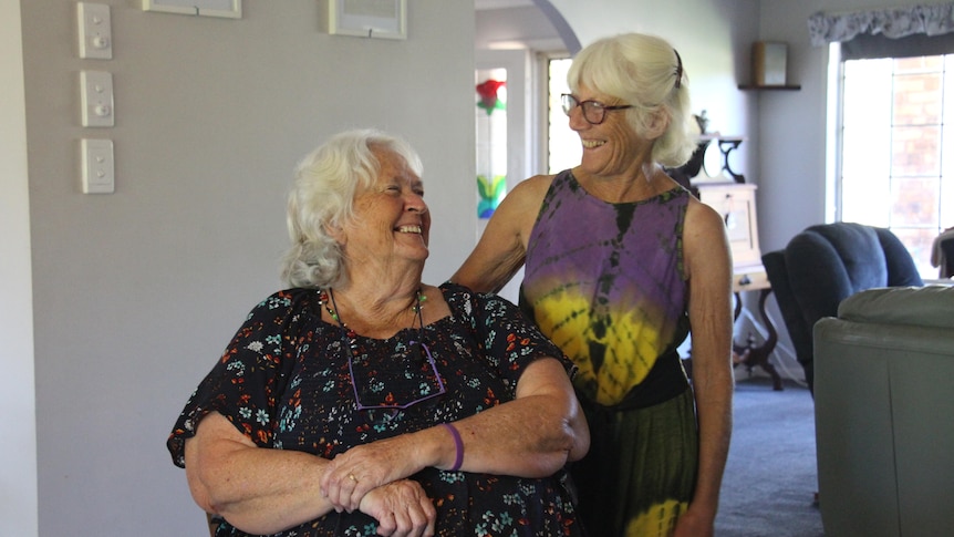 Two women with white hair, one sitting down, look at each other smiling with a living room behind them