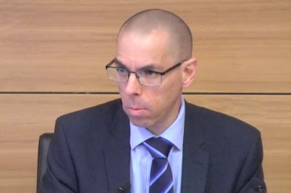 A man with glasses and a suit in front of a wooden panel.