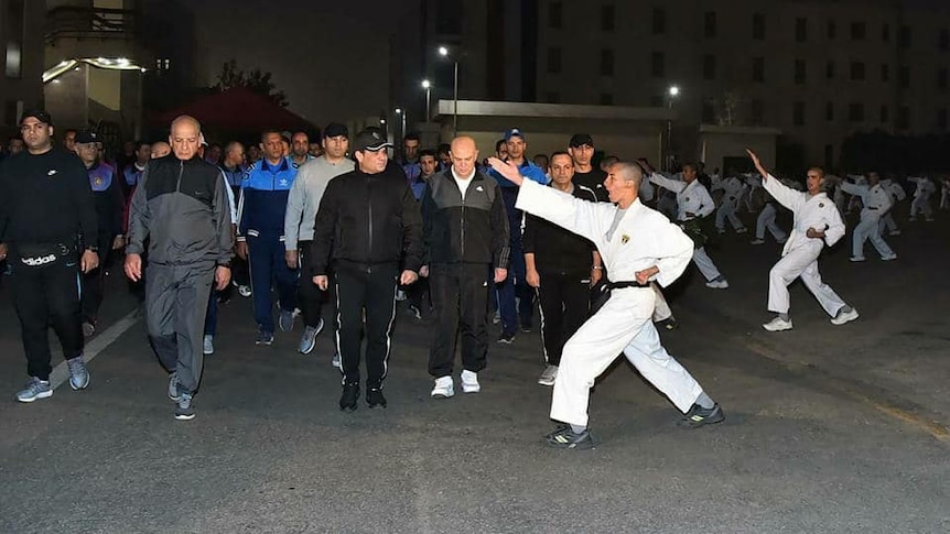 Egyptian President el Sissi walks in activewear down a nighttime street, viewing black-belt karate performers mid-salute.