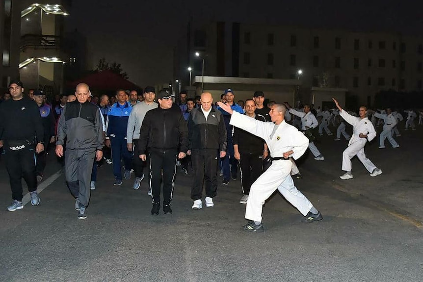 Egyptian President el Sissi walks in activewear down a nighttime street, viewing black-belt karate performers mid-salute.