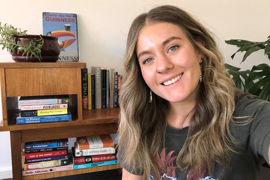 A young woman with blonde hair smiles, behind her is a bookcase 