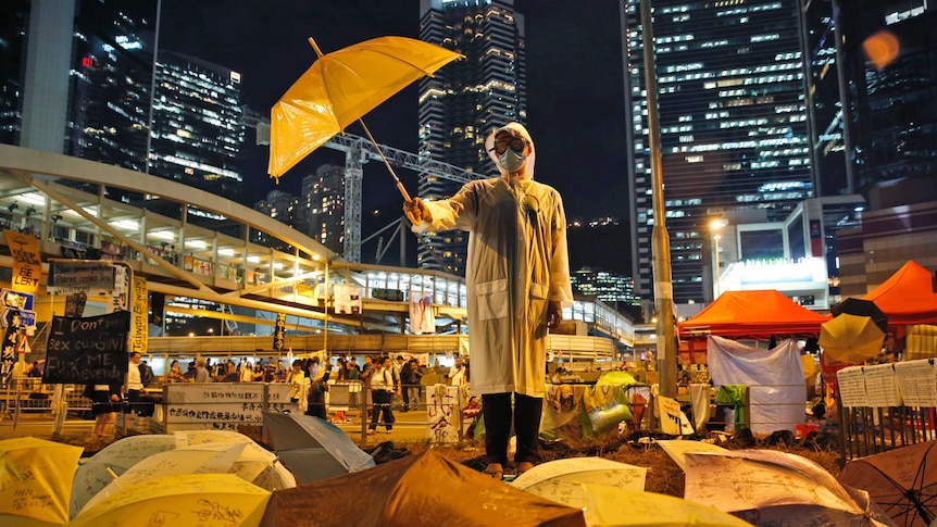 A Hong Kong protester wears a face mask and holds an umbrella defiantly in front of high-rise buildings