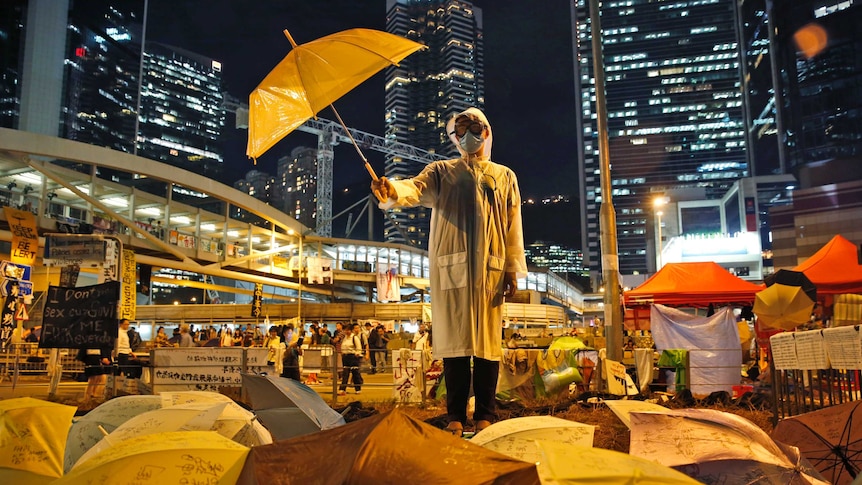 A Hong Kong protester wears a face mask and holds an umbrella defiantly in front of high-rise buildings