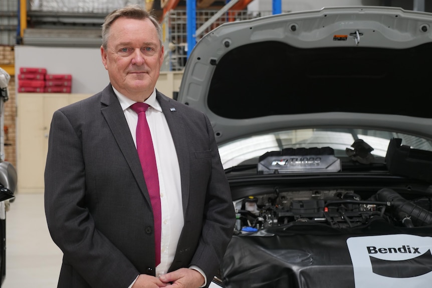 A man in a suit and tie stands in front of a car with the bonnet open