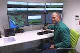 A man sits in front of a desk with six computer screens. They show various views of plantation forests.