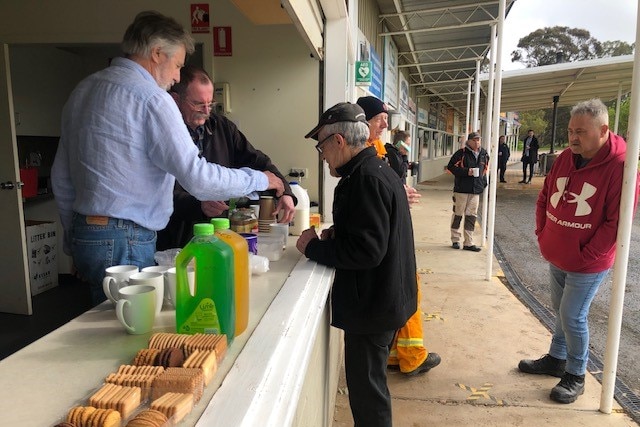 Two men serving another man over a counter with biscuits and drinks