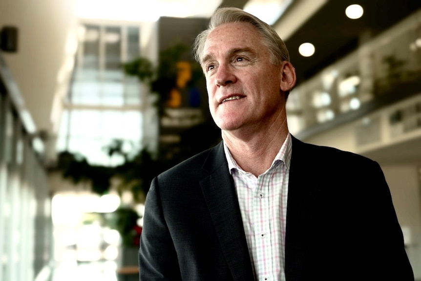 A grey-haired man in a suit jacket and checked shirt looks to his right, off camera, as he stands in the lobby of a building