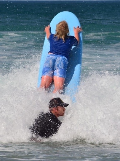 A man in a black top with a black hat in the surf with a boy on a blue surfboard.