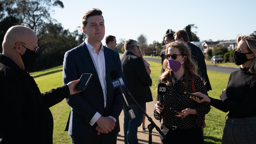 Young man stands in front of reporter mics on sunny day, reporters wearing face masks