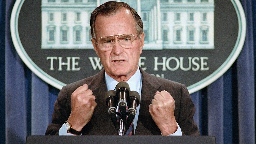 a man with glasses stands at a lectern with his hands in fists