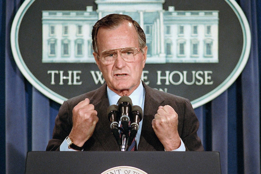 a man with glasses stands at a lectern with his hands in fists