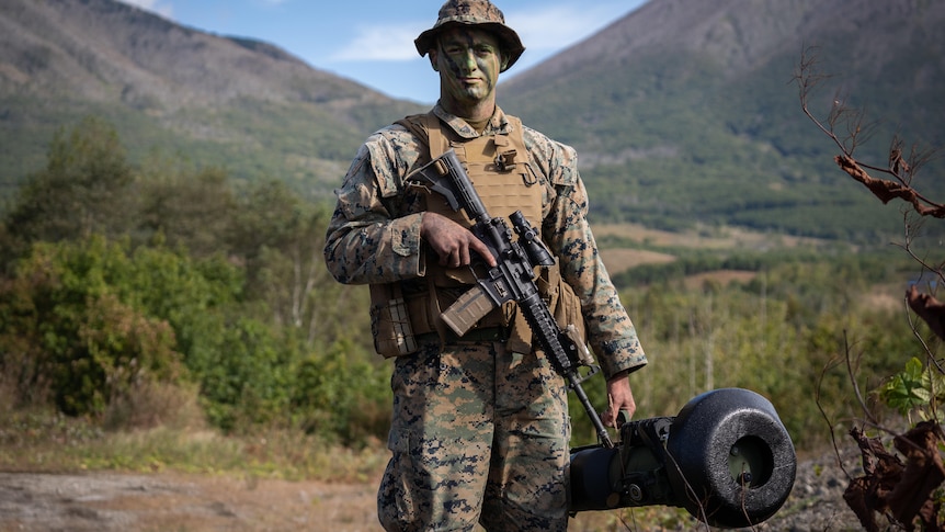 A soldier in camo gear with a rifle across his chest carries a huge black missile system