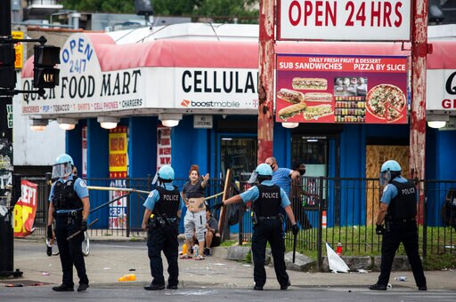 Four police officers in face shields stand in front of a shop, a woman is gesturing to the officers