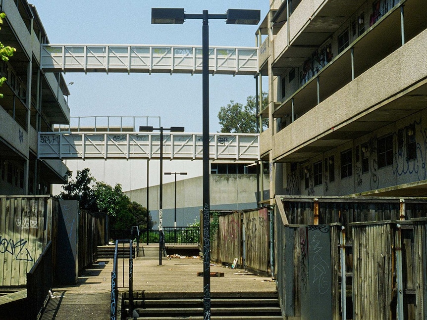 On a blue day, you view an abandoned public housing estate with boarded up windows and graffiti.