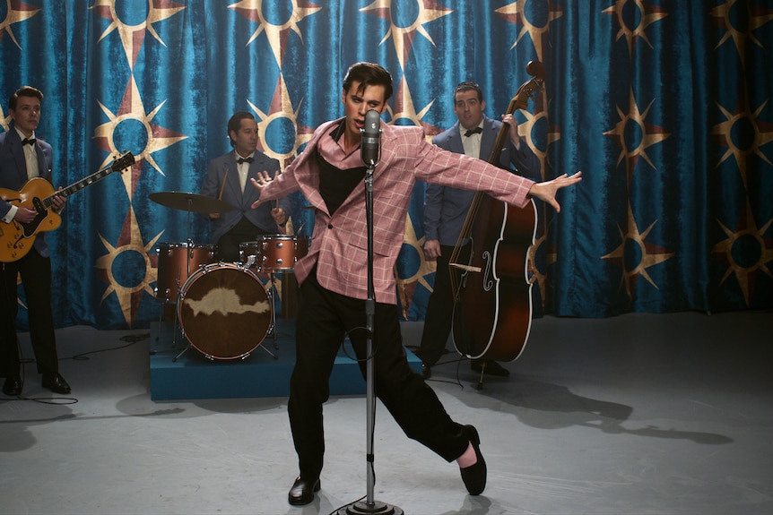 Un homme blanc habillé en Elvis avec une veste rose et les cheveux lissés, se produit sur scène avec un groupe et prend des poses.