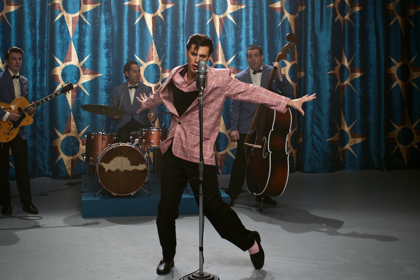 Un homme blanc habillé en Elvis dans un blazer rose et aux cheveux ébouriffés se produit sur scène avec un groupe et prend la pose.